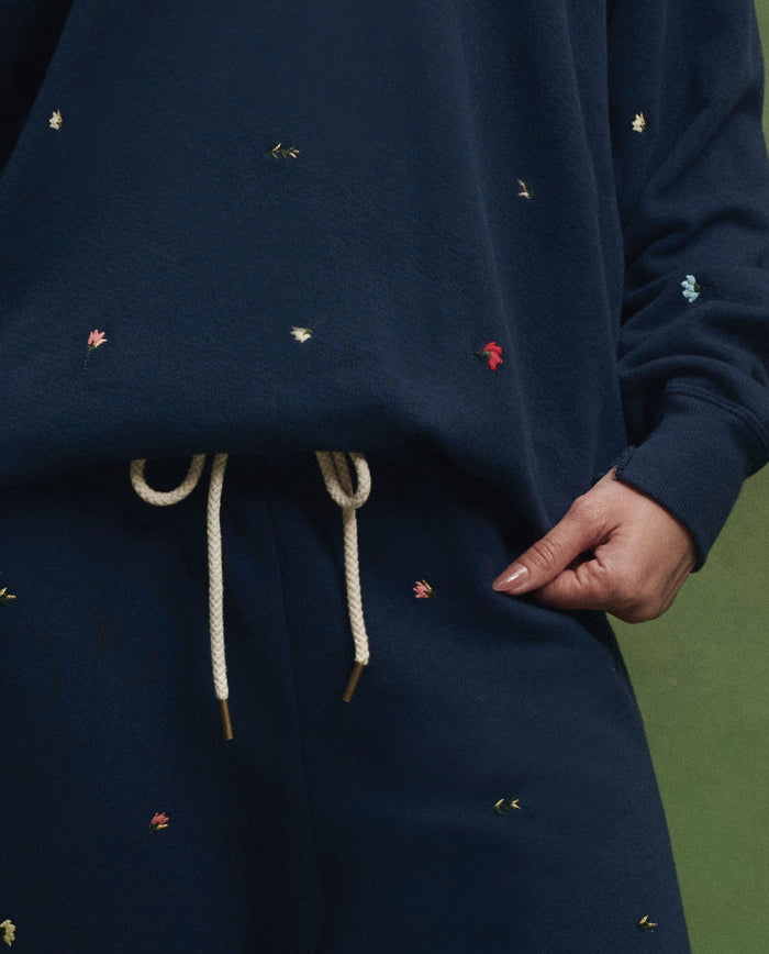 A person models THE GREAT. PS26’s The Stadium Sweatpant. Embroidered—Navy with Tossed Floral Embroidery—featuring small colorful florals and beige drawstrings, evoking vintage athletic style against a green backdrop.