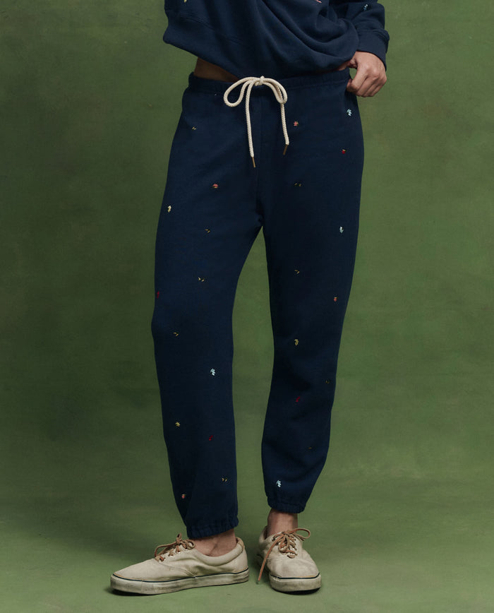 Someone wears THE GREAT. PS26’s The Stadium Sweatpant. Embroidered—navy with tossed floral embroidery—paired with a matching vintage-inspired top and beige lace-up shoes, standing before a green backdrop.