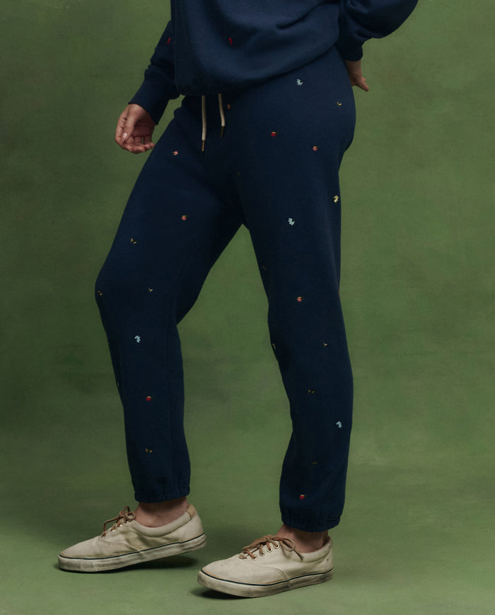 Model wears THE GREAT. PS26 The Stadium Sweatpant in navy with tossed floral embroidery, paired with beige sneakers, standing before a green background. Inspired by vintage athletic wear and detailed with delicate embroidered florals.