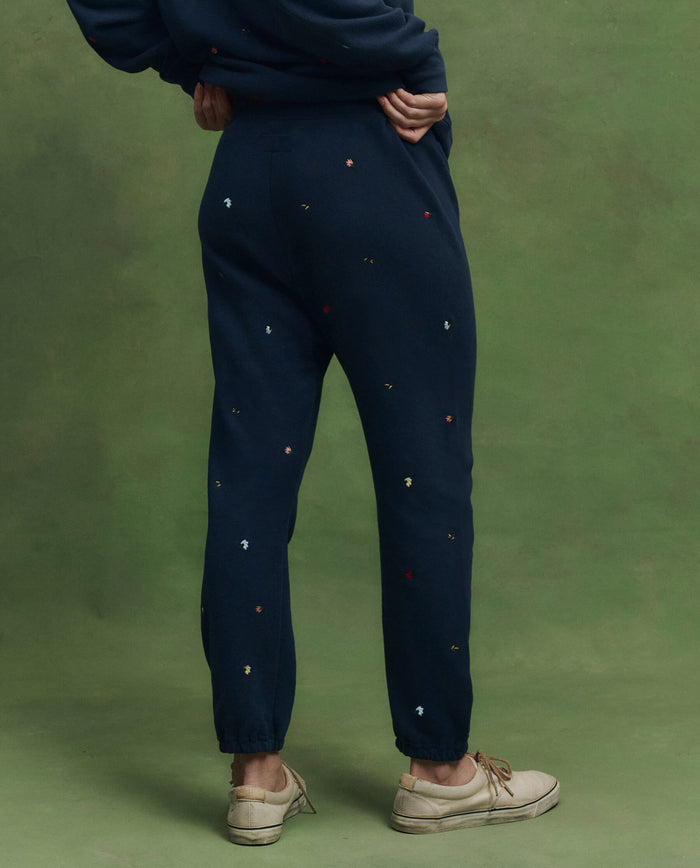 A person stands on a green background wearing THE GREAT. PS26 The Stadium Sweatpant. Embroidered—navy with tossed floral embroidery—paired with a matching sweatshirt and beige lace-up shoes; classic vintage athletic wear made in Los Angeles.