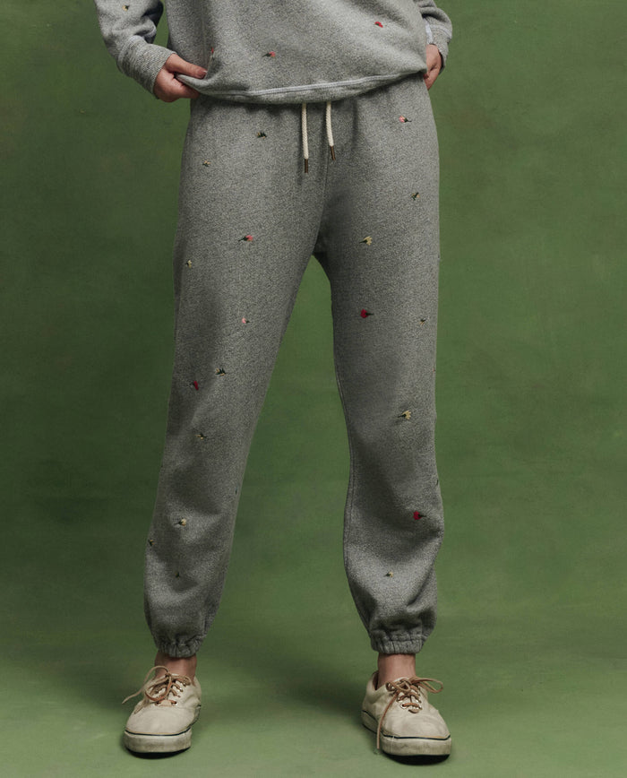 A person wears THE GREAT. SU26 The Stadium Sweatpant W/ Tossed Floral Emb. in Varsity Grey, featuring small floral embossing, shown from the waist down with white sneakers against a green background.