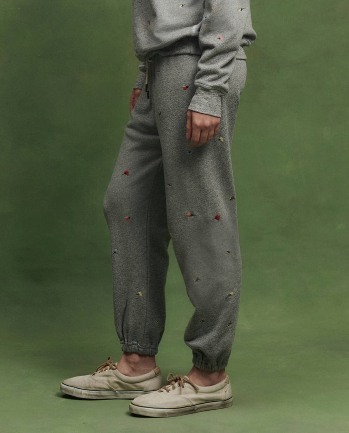 A person stands against a green backdrop wearing THE GREAT. SU26 The Stadium Sweatpant W/ Tossed Floral Emb. in Varsity Grey, paired with a matching sweatshirt and white sneakers.