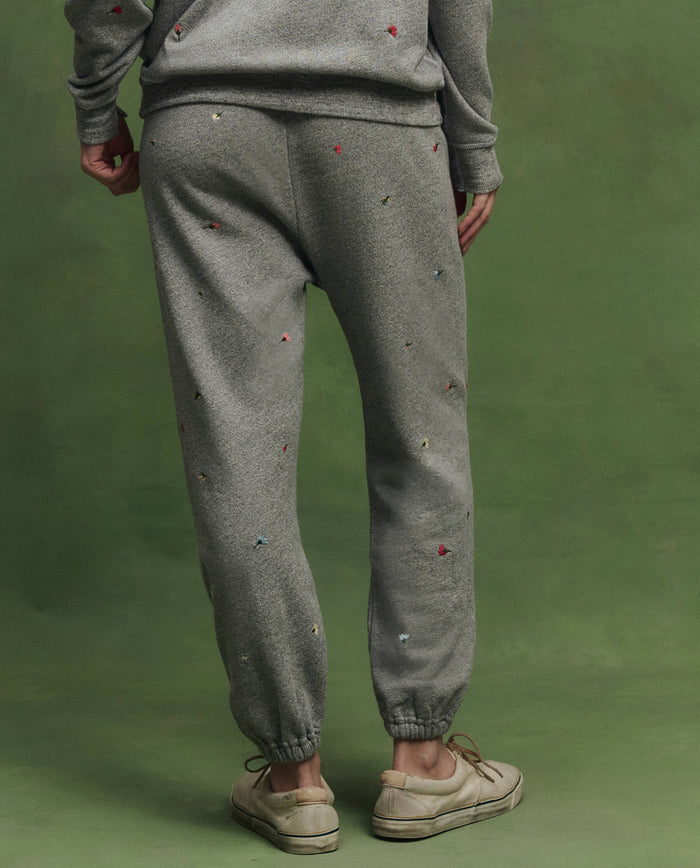 A person stands against a green background wearing THE GREAT. SU26 The Stadium Sweatpant W/ Tossed Floral Emb. in Varsity Grey, paired with a matching sweatshirt featuring colorful floral embroidery and beige sneakers.