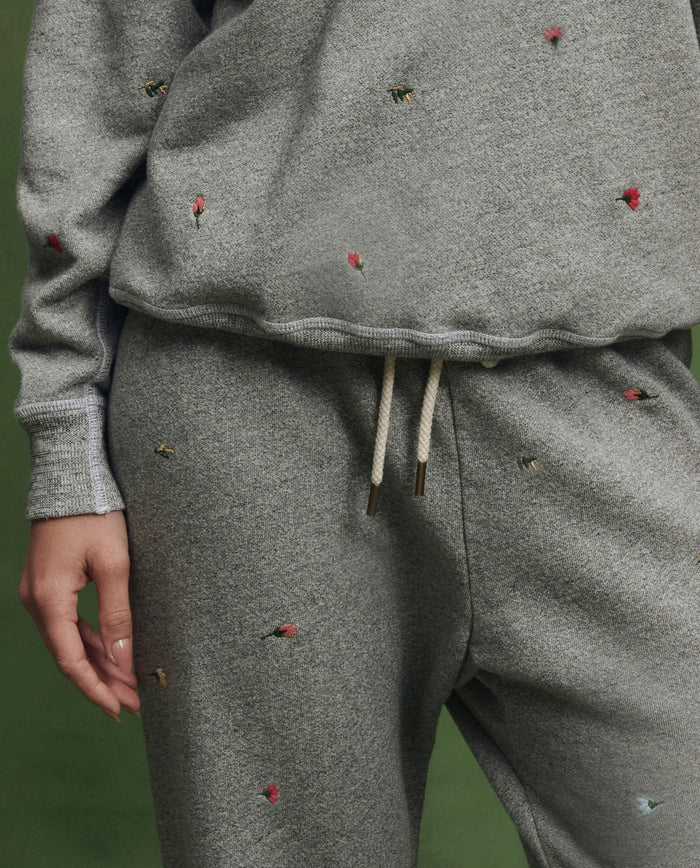 Model wearing THE GREAT. SU26's The Stadium Sweatpant W/ Tossed Floral Emb. in Varsity Grey, shown from shoulders to thighs, against a green background.