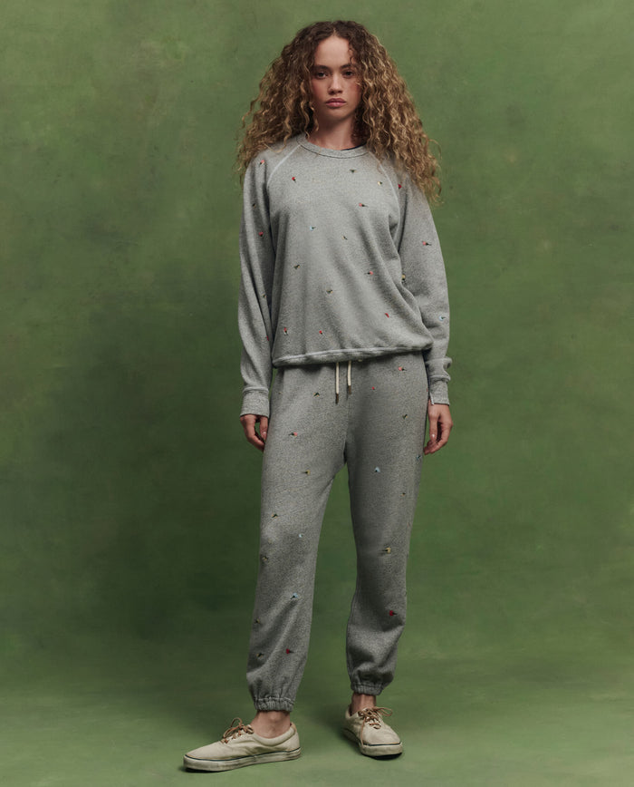 A woman with curly hair stands against a green background, wearing THE GREAT. SU26's The Stadium Sweatpant W/ Tossed Floral Emb. in Varsity Grey, paired with a matching sweatshirt and beige sneakers.