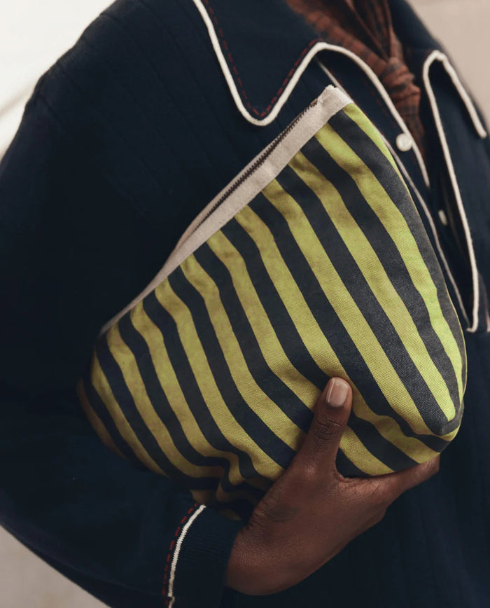 A person holds THE GREAT. F26 D1 The Stripe Pouch in Citron Mini Prep Stripe—a green and black striped clutch bag crafted from cotton canvas—while wearing a dark shirt with white piping on the collar.