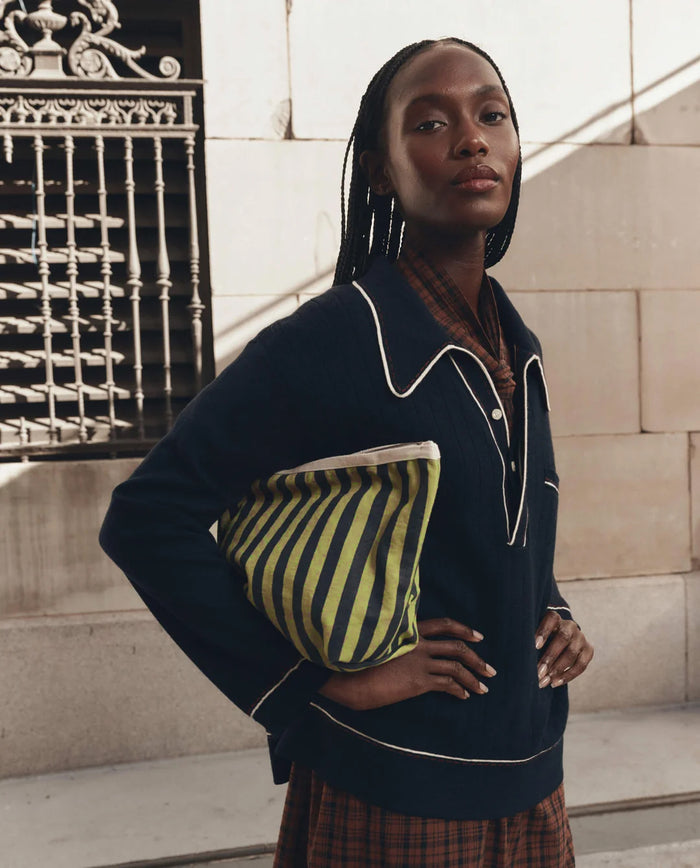 A woman in a navy sweater with white trim stands outdoors near a stone building with a black iron fence, holding THE GREAT. F26 D1′s The Stripe Pouch in Citron Mini Prep Stripe—the perfect travel companion for her day out.