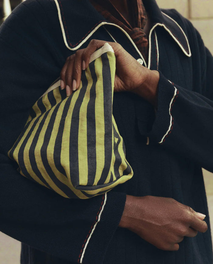 Carrying THE GREAT. F26 D1′s “The Stripe Pouch” in Citron Mini Prep Stripe—a soft cotton canvas bag featuring green and black vertical stripes—this person, dressed in a navy sweater with white-trimmed cuffs and collar, has a stylish travel companion for any journey.