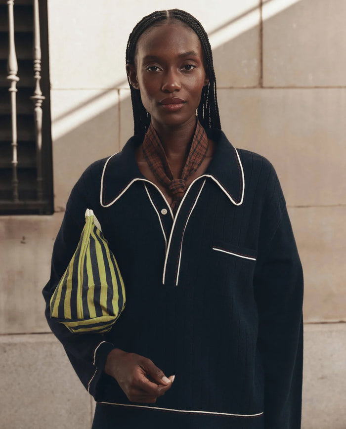 A woman with braided hair stands against a stone wall, wearing a dark collared sweater and a brown scarf, while holding THE GREAT. F26 D1 Stripe Pouch in Citron Mini Prep Stripe, crafted from cotton canvas.