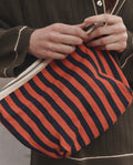 A person holding THE GREAT. F26 D1 The Stripe Pouch in Hot Red Mini Prep Stripe, styled with a dark long-sleeve top featuring white trim.