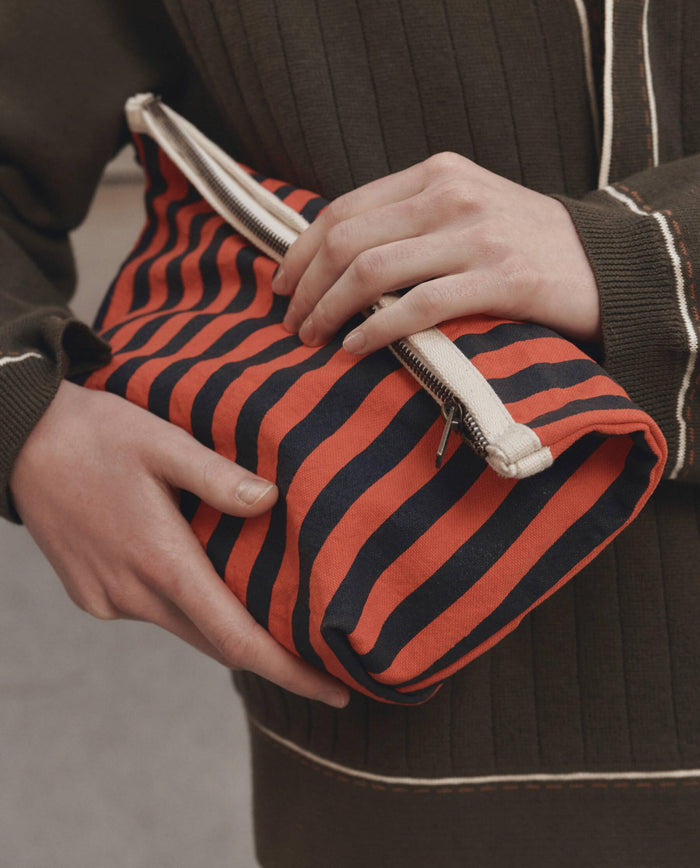 A person wearing a dark green sweater holds The Stripe Pouch in Hot Red Mini Prep Stripe by THE GREAT. F26 D1, featuring stylish diagonal red and navy stripes and a white zipper.