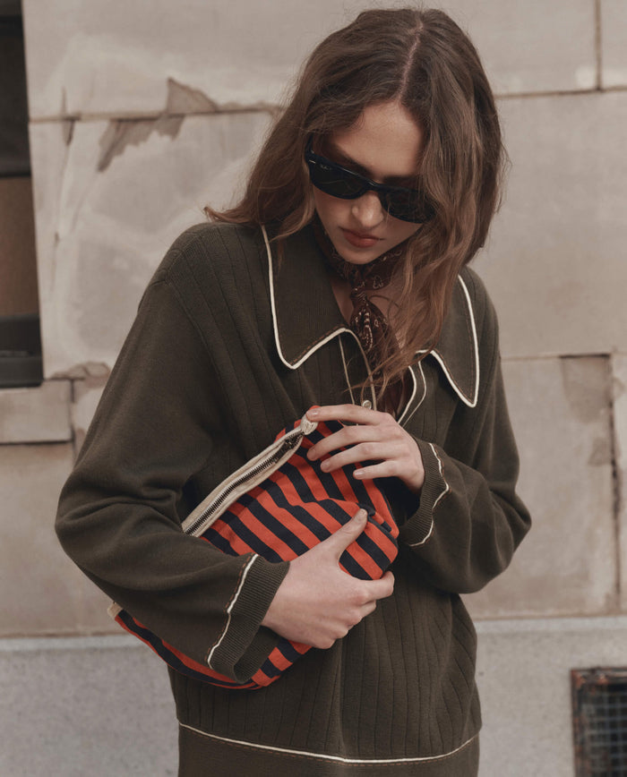 A woman with wavy hair wears black sunglasses, a patterned neckerchief, and an olive green knit shirt with contrast piping, holding THE GREAT. F26 D1's The Stripe Pouch in Hot Red Mini Prep Stripe in front of her.