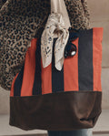 A person carries THE GREAT. F26 D1 "The Stripe Tote" in Hot Red Prep Stripe, featuring bold red and black stripes, a brown leather bottom, a knotted bandana, and a pair of sunglasses attached to the handle.