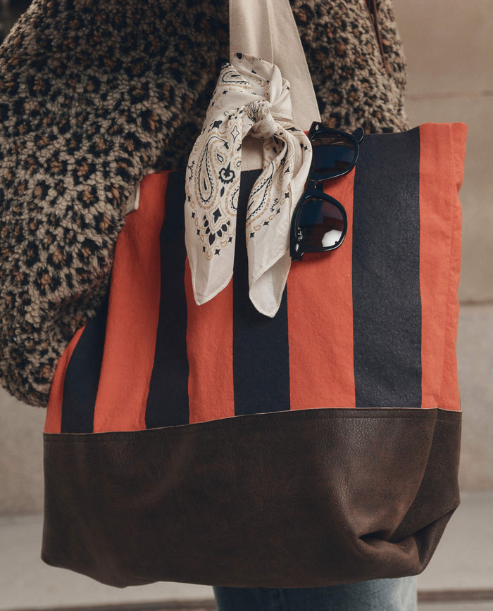 A person carries THE GREAT. F26 D1 "The Stripe Tote" in Hot Red Prep Stripe, featuring bold red and black stripes, a brown leather bottom, a knotted bandana, and a pair of sunglasses attached to the handle.