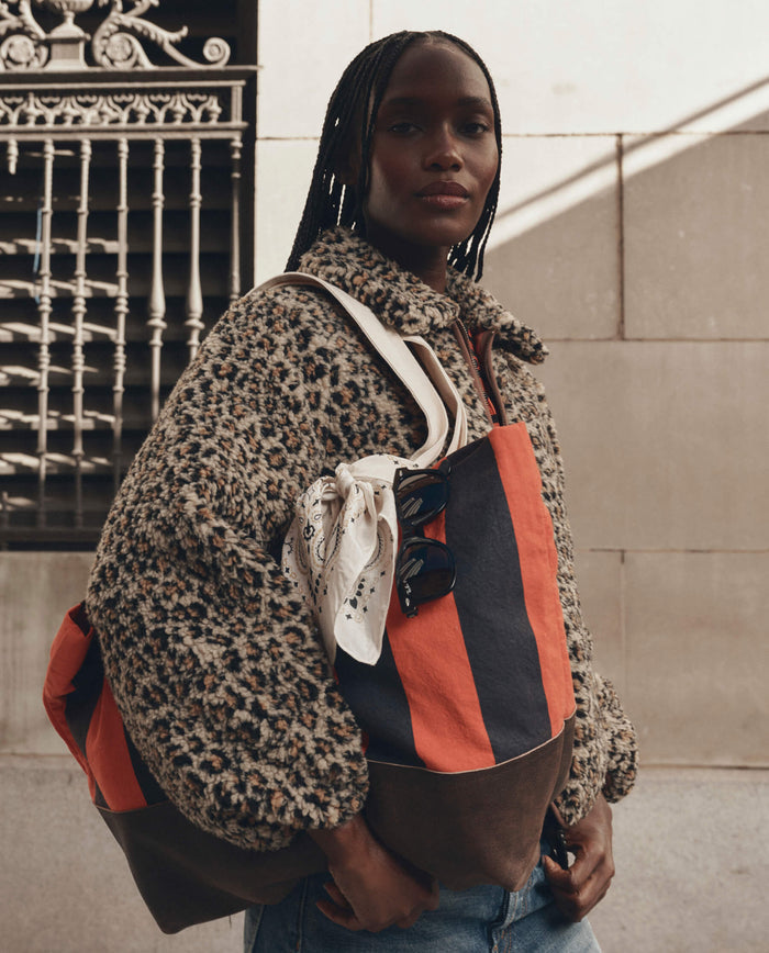 A person wearing a leopard print jacket and jeans carries THE GREAT. F26 D1 The Stripe Tote in Hot Red Prep Stripe, accented with sunglasses and a bandana attached to the handles, standing in front of a building.