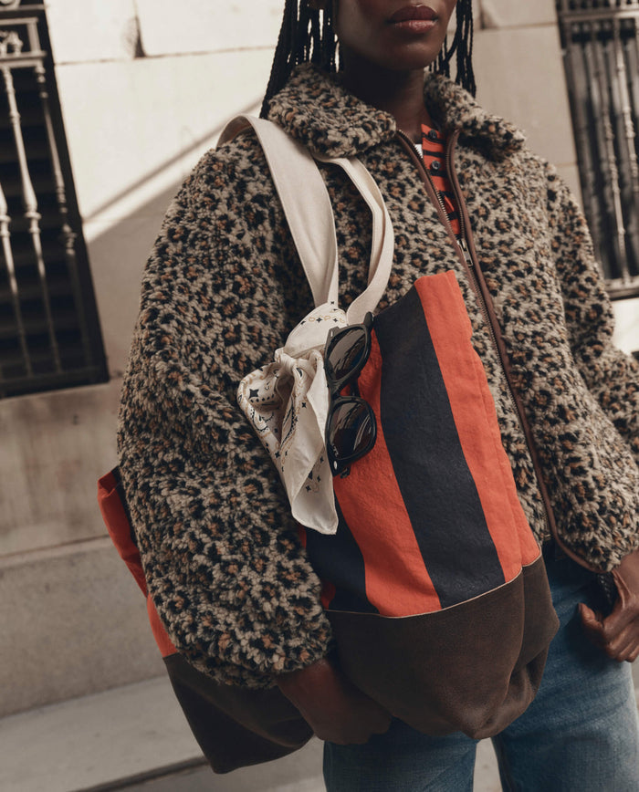 A person wearing a faux fur leopard print jacket carries two tote bags, one of which is THE GREAT.'s The Stripe Tote in Hot Red Prep Stripe, and also wears a white scarf with sunglasses attached while standing outdoors.