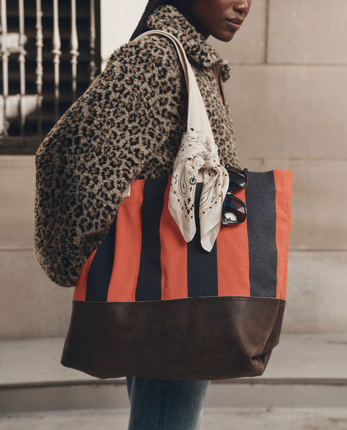 A person wearing a leopard-print fleece jacket carries THE GREAT. F26 D1 The Stripe Tote in Hot Red Prep Stripe—a canvas tote bag with orange and black stripes, brown leather trim, a knotted bandana on the strap, and sunglasses tucked in.