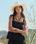 A woman stands outdoors in the Structured Victorian Tank by THE GREAT. PF26 in black, paired with a white cowboy hat, beaded necklace, and bracelets; desert plants and hills provide a natural backdrop.