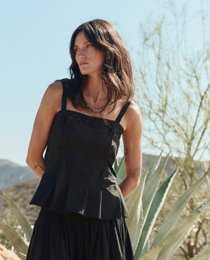 A woman with long dark hair wears THE GREAT. PF26 The Structured Victorian Tank in black, standing outdoors in a desert landscape with cacti and dry plants in the background.