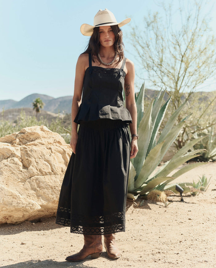 A woman stands outdoors in a desert landscape wearing THE GREAT. PF26 The Structured Victorian Tank in black, paired with a white cowboy hat and long black skirt, surrounded by rocks, cacti, and mountains.