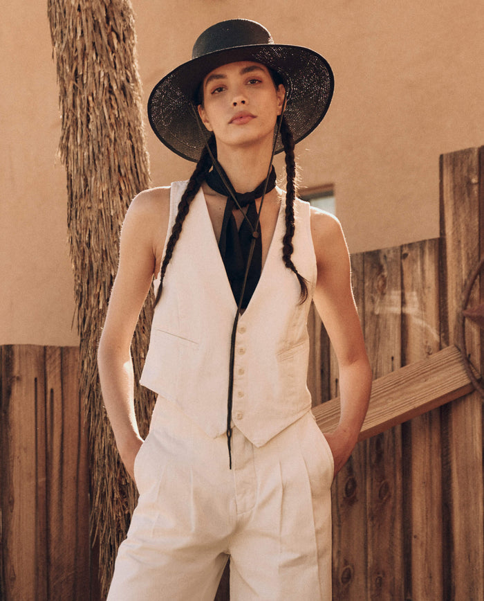 A person wearing a light-colored suit vest by THE GREAT. PF24 in natural hemp, paired with a black hat and black necktie, stands outdoors near a wooden fence.