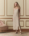 A woman in THE GREAT. SP26 D2 The Sunroom Slip Dress—Champagne Scattered Bloom—stands beside a pink upholstered sofa, against beige paneled walls, wearing brown oxford shoes.