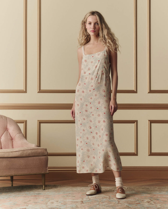 A woman in THE GREAT. SP26 D2 The Sunroom Slip Dress—Champagne Scattered Bloom—stands beside a pink upholstered sofa, against beige paneled walls, wearing brown oxford shoes.