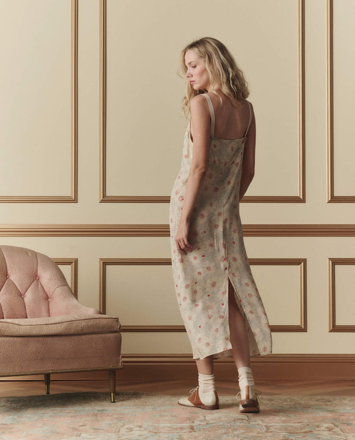 A woman wearing THE GREAT. SP26 D2 The Sunroom Slip Dress in Champagne Scattered Bloom stands with her back to the camera in a beige-paneled room with a pink upholstered chair.