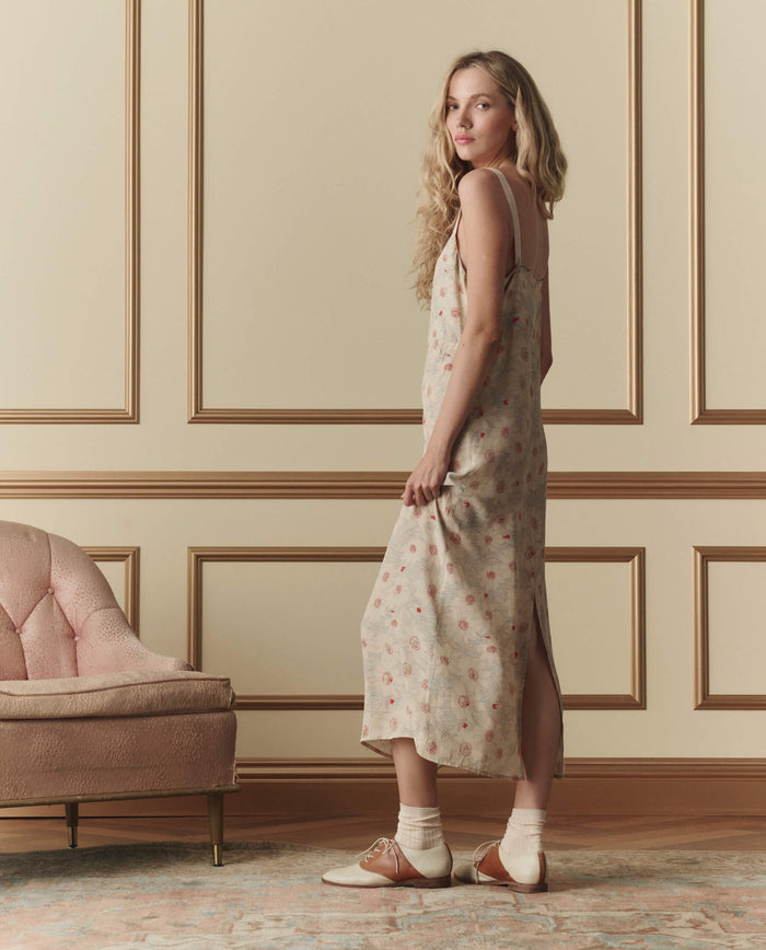 A woman in THE GREAT. SP26 D2 The Sunroom Slip Dress—Champagne Scattered Bloom stands sideways by a pink upholstered chair in a paneled room, wearing oxford shoes with socks.
