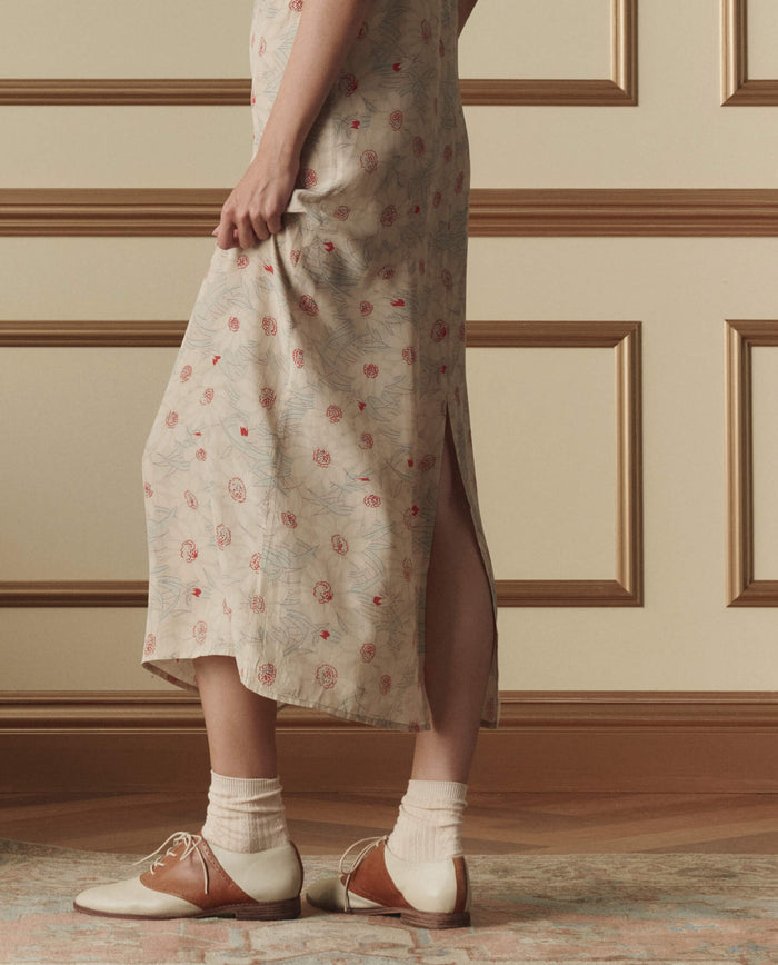 A person wearing THE GREAT. SP26 D2 The Sunroom Slip Dress in Champagne Scattered Bloom, cream socks, and two-tone brown and cream oxford shoes stands indoors on a decorative rug.