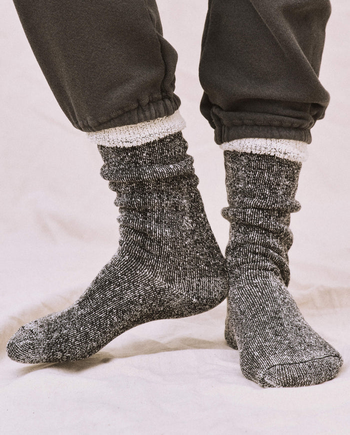 A person stands on a light-colored surface wearing black THE GREAT. SP25 SWEATSHIRT SOCKS, a cozy sweatshirt, and dark pants with elastic cuffs.