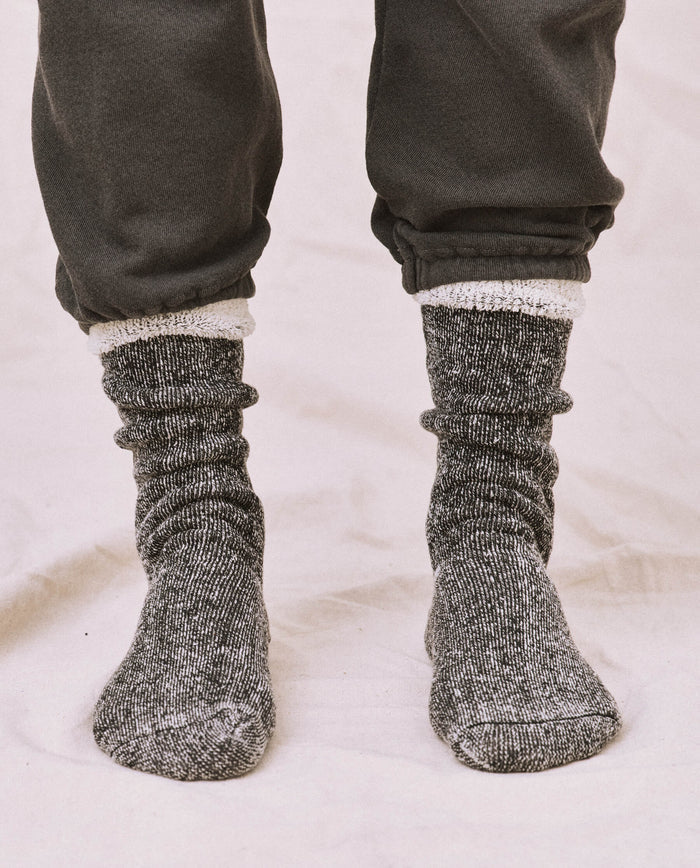 A person wears dark pants and a cozy sweatshirt with THE GREAT. SP25 Sweatshirt Socks in black, their pants cuffed to reveal white sock liners against a light background.