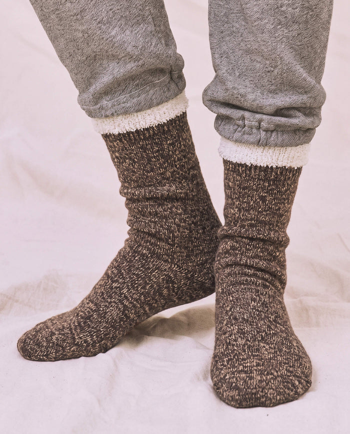 Wearing THE GREAT. CARRYOVER The Sweatshirt Sock in Chocolate with gray sweatpants, standing on a light-colored surface.