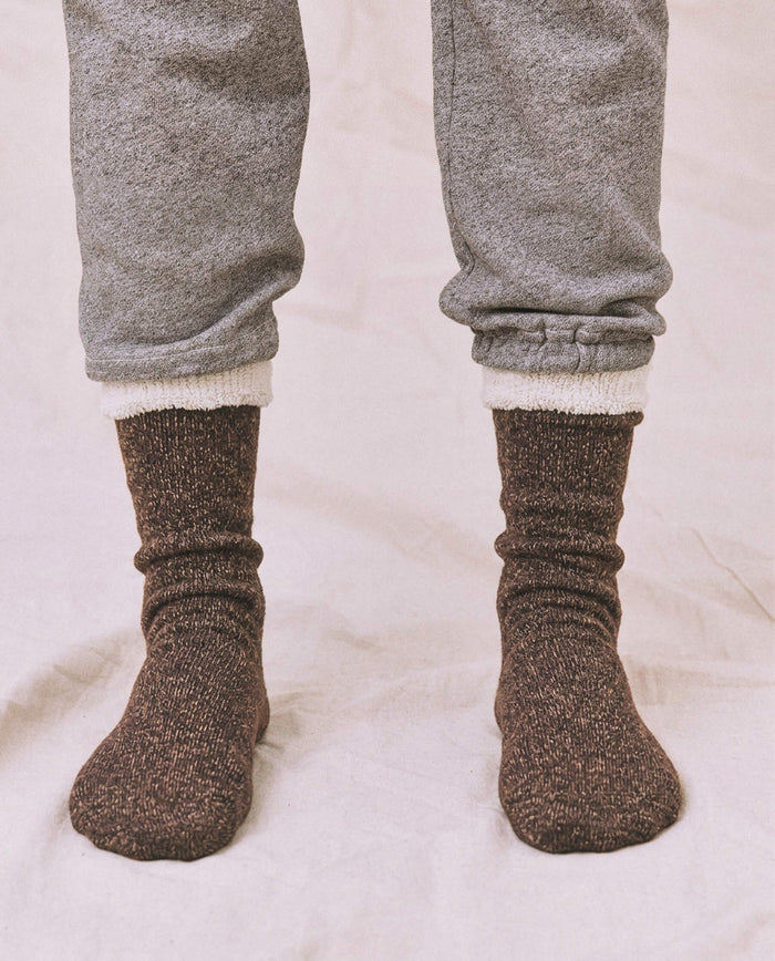 A person wearing THE GREAT. CARRYOVER The Sweatshirt Sock in Chocolate stands on a light-colored surface, with their grey sweatpants slightly rolled up above the ankles.