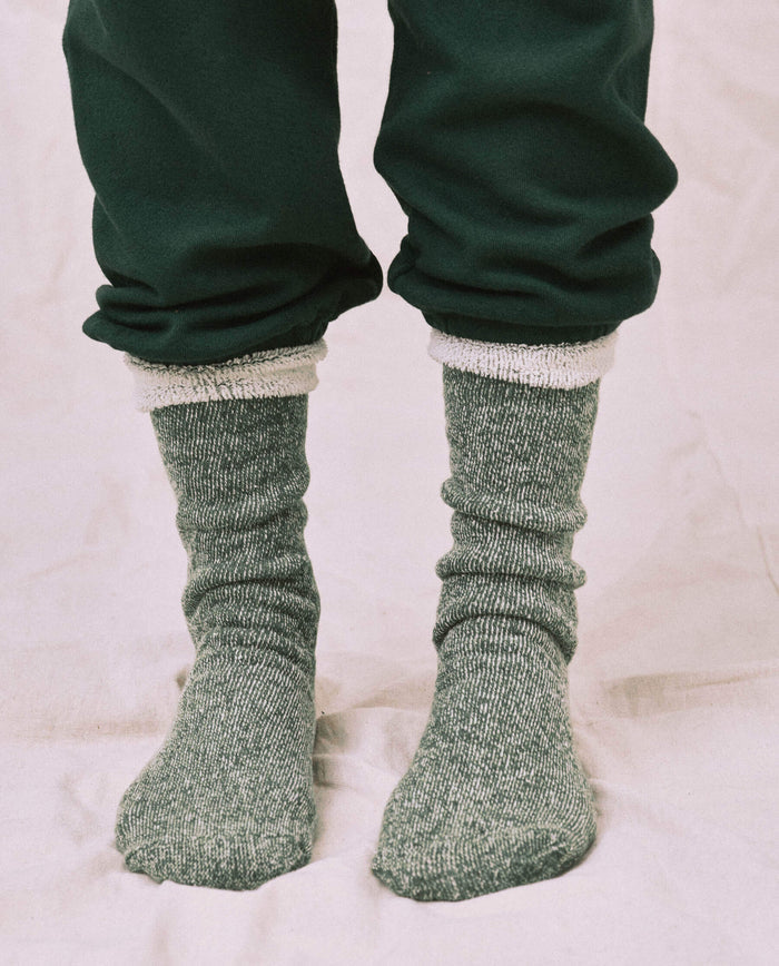 Someone wearing THE GREAT. CARRYOVER The Sweatshirt Sock. -- Deep Pine stands on a light-colored fabric surface, paired with dark green pants and showcasing the cozy, thick style of the socks.