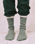 Someone wearing THE GREAT. CARRYOVER The Sweatshirt Sock. -- Deep Pine stands on a light-colored fabric surface, paired with dark green pants and showcasing the cozy, thick style of the socks.
