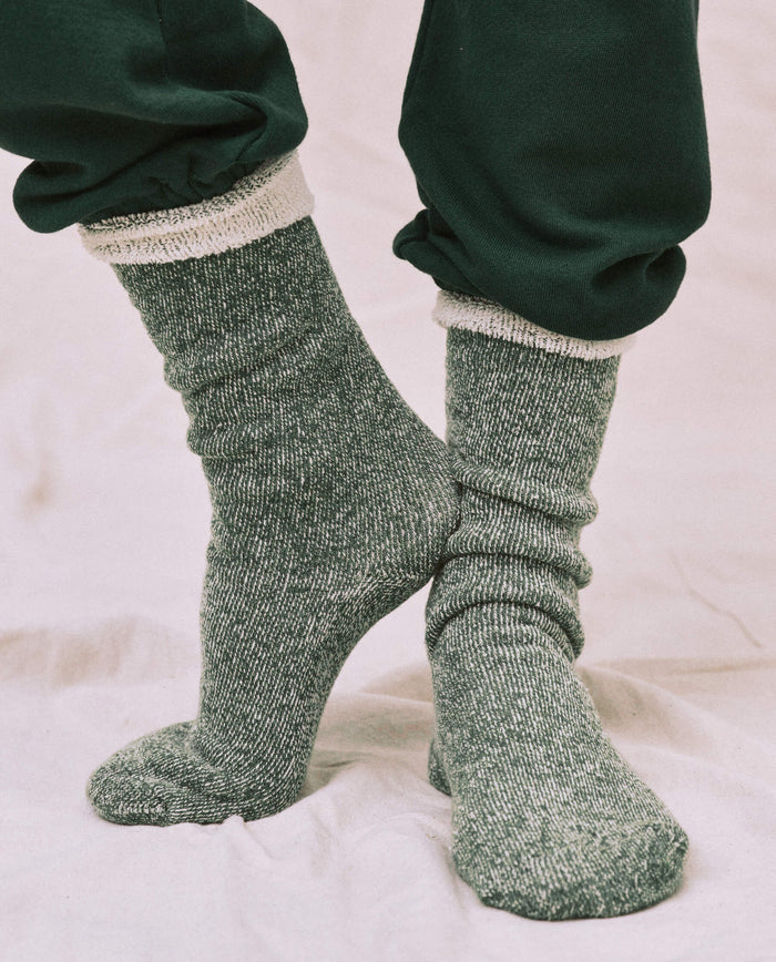 Someone wearing THE GREAT. CARRYOVER's Deep Pine Sweatshirt Socks, featuring a green and white speckled design, stands on a light-colored surface in dark pants.
