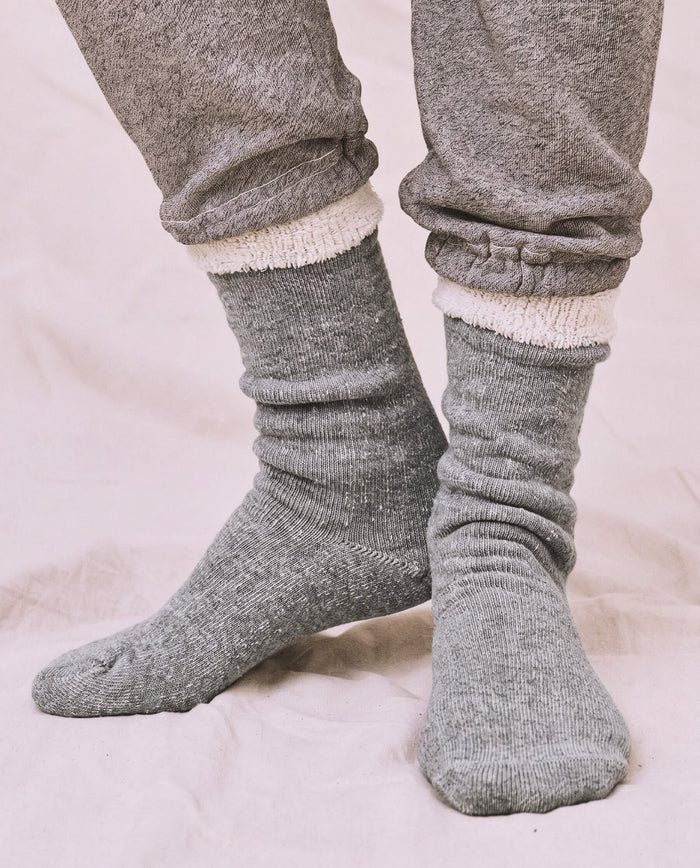 A person wearing thick, heather grey The Sweatshirt Sock by THE GREAT. CARRYOVER stands on a light-colored fabric surface, paired with gray French Terry sweatpants.
