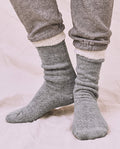 A person wearing thick, heather grey The Sweatshirt Sock by THE GREAT. CARRYOVER stands on a light-colored fabric surface, paired with gray French Terry sweatpants.