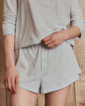 A person wearing THE GREAT. H25 SLEEP Tap Short in Heather Grey with a gathered waistband and a light grey long-sleeve top, standing against a wooden background—ideal for cozy nights.