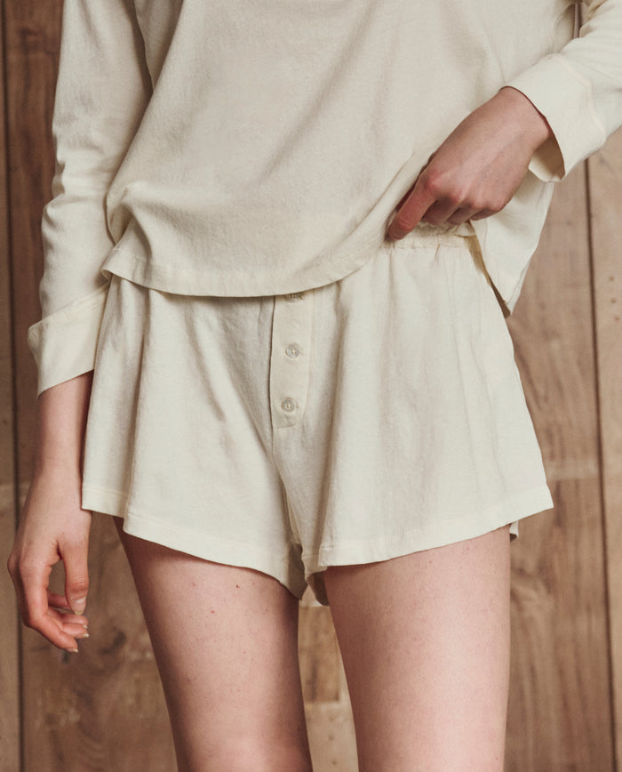A person wearing THE GREAT. H25 SLEEP’s The Tap Short in Washed White, a loose 100% cotton sleep short, paired with a matching long-sleeve top, stands against a wooden background.