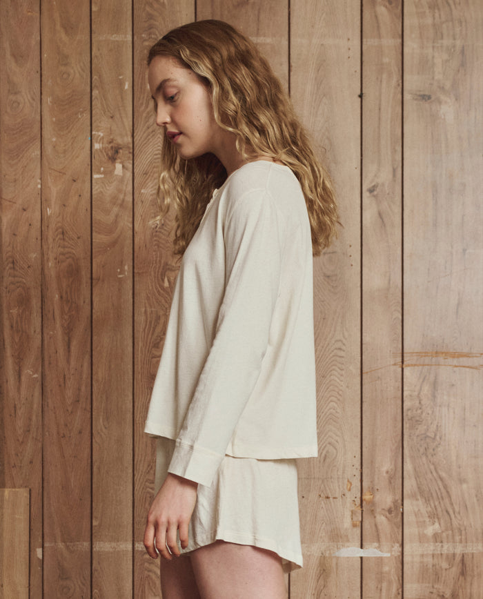 A woman with wavy blonde hair stands in profile against a wooden wall, wearing a loose beige top and THE GREAT. H25 SLEEP The Tap Short in Washed White, made from 100% cotton.