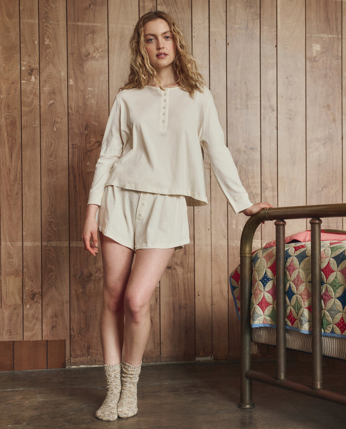 A person stands indoors by a colorful quilted bed, wearing THE GREAT. H25 SLEEP’s The Tap Short.. in Washed White and a cream long-sleeve top, with a wooden wall in the background.