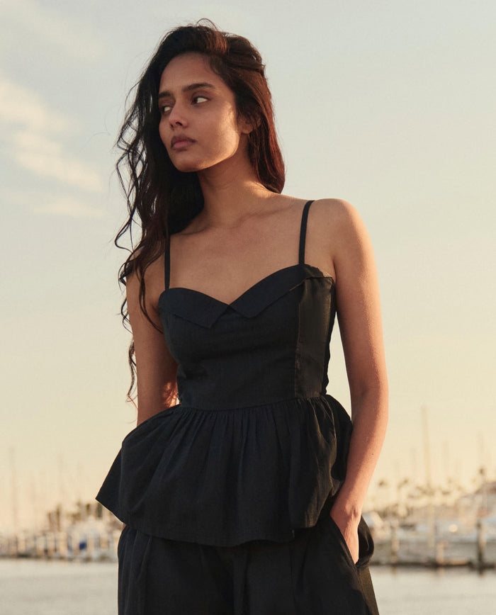 A woman with long dark hair wears THE GREAT. SU26 The Verse Top in black while standing outdoors near a marina, boats and masts visible behind her.
