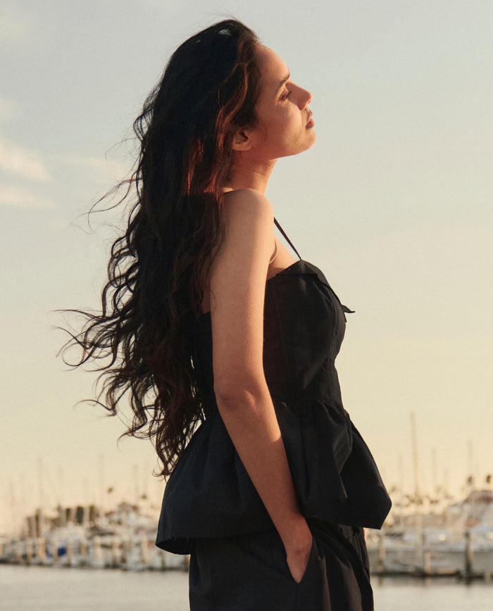 A woman with long wavy hair stands outdoors near a marina at sunset, facing sideways with hands in pockets, wearing THE GREAT. SU26 The Verse Top in black.