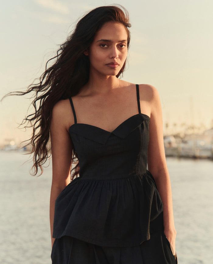 A woman with long dark hair stands outdoors near water, wearing THE GREAT. SU26 The Verse Top in black, with a marina and boats visible in the background.