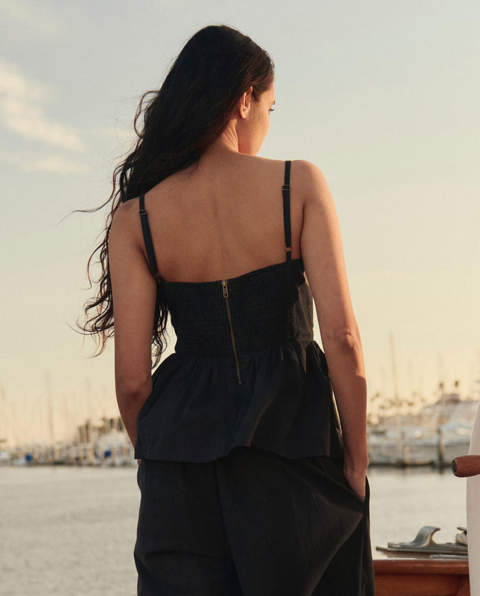 A woman with long dark hair stands on a dock facing away, wearing THE GREAT. SU26 The Verse Top in black and matching pants, with boats and water in the background.