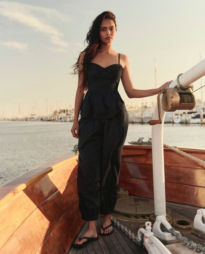 A woman in THE GREAT. SU26 The Verse Top – Black, black pants, and sandals stands on a wooden boat by the water with a marina and boats in the background.