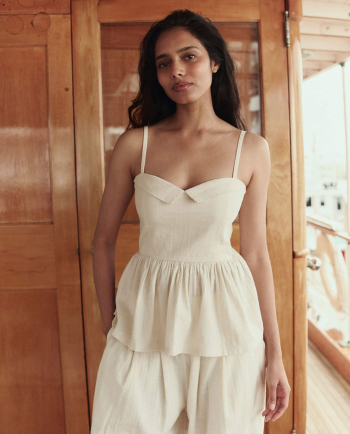 A woman stands indoors wearing THE GREAT. SU26's The Verse Top in Natural, a cream-toned piece with thin straps, paired with matching loose pants against a wooden backdrop.