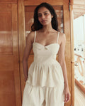 A woman stands indoors wearing THE GREAT. SU26's The Verse Top in Natural, a cream-toned piece with thin straps, paired with matching loose pants against a wooden backdrop.